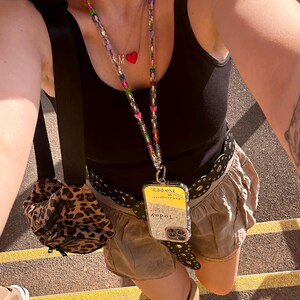 May include: A black tank top, tan shorts, and a black belt with a decorative design. A phone case with the word "Today" and "Happy" is attached to a beaded lanyard. A small leopard print bag is visible.