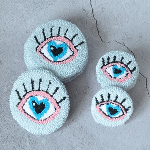 May include: Four blue and pink eye-shaped poufs with black eyelashes and a heart-shaped pupil. The poufs are made of yarn and are arranged in a circle on a gray background.