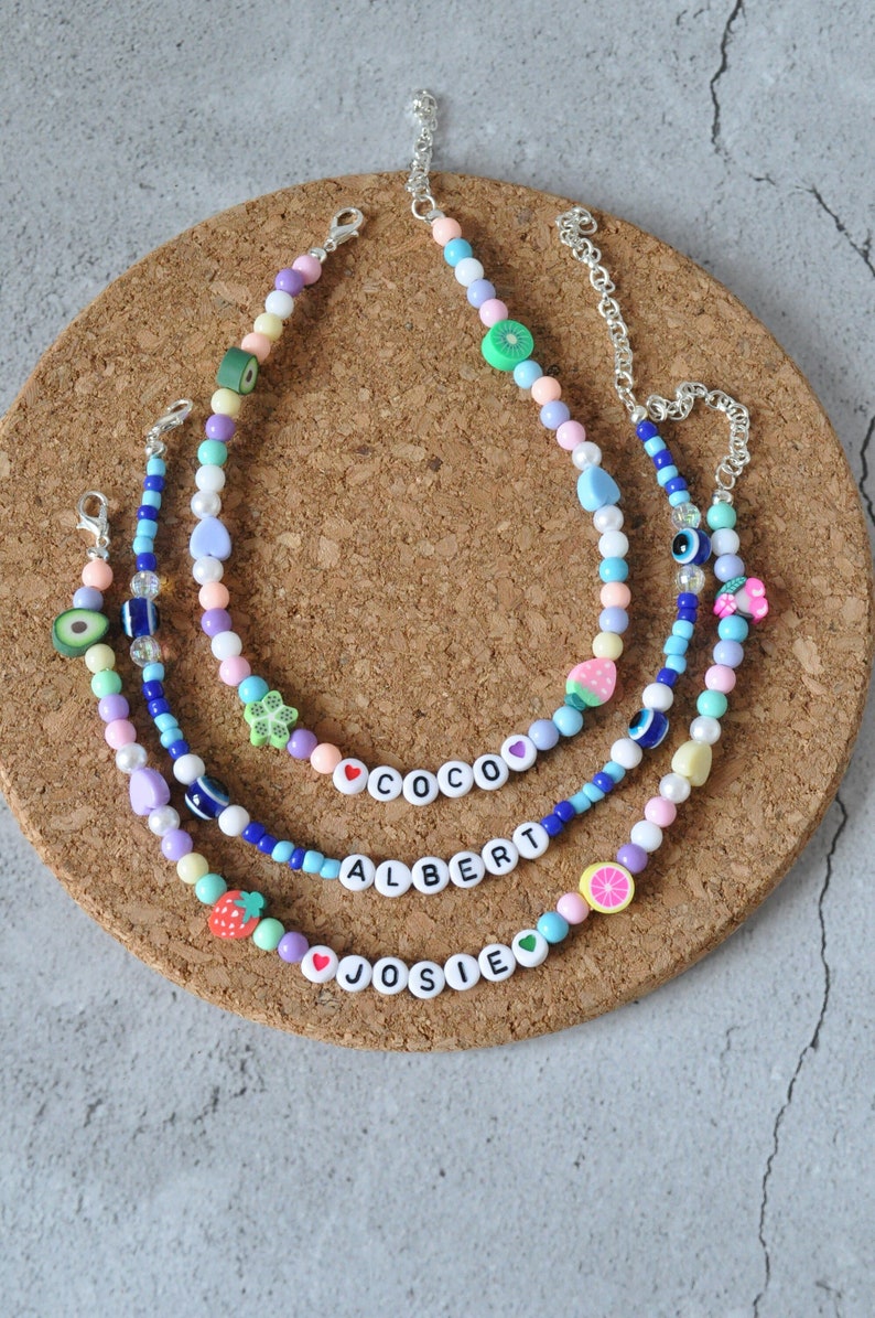 May include: Three colorful beaded necklaces with charms, including fruit, hearts, and evil eyes. The necklaces spell out the names "Coco", "Albert", and "Josie".