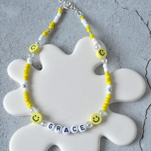 May include: A yellow and white beaded necklace with smiley face charms and white letter beads spelling out the name "GRACE".