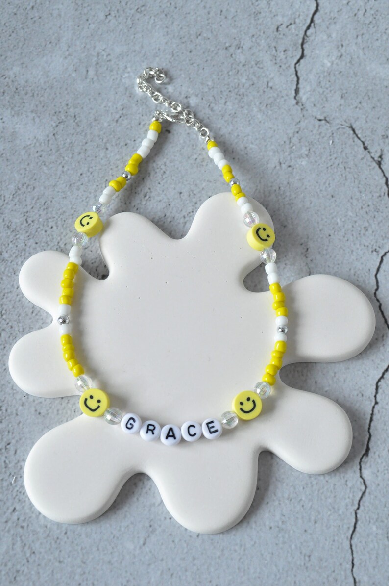 May include: A yellow and white beaded necklace with smiley face charms and white letter beads spelling out the name "GRACE".