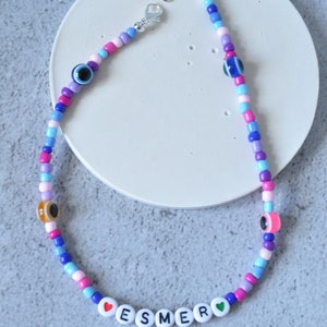 May include: A colorful beaded necklace with a silver clasp. The necklace features blue, pink, purple, and white beads, as well as small evil eye charms. The necklace also has white letter beads that spell out "ESMER" with a red heart on the left and a green heart on the right.