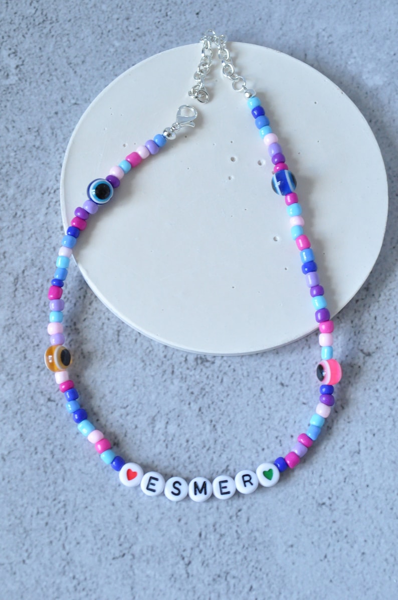 May include: A colorful beaded necklace with a silver clasp. The necklace features blue, pink, purple, and white beads, as well as small evil eye charms. The necklace also has white letter beads that spell out "ESMER" with a red heart on the left and a green heart on the right.