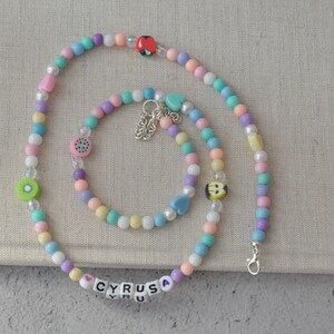 May include: A colorful beaded necklace and bracelet set with a heart charm and the name "CYRUS" spelled out in white beads. The necklace and bracelet are made of pastel pink, blue, green, and yellow beads.