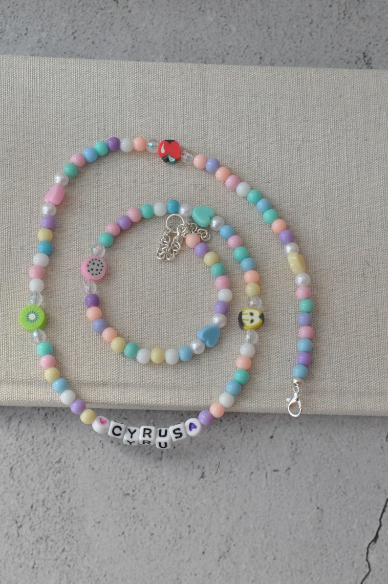 May include: A colorful beaded necklace and bracelet set with a heart charm and the name "CYRUS" spelled out in white beads. The necklace and bracelet are made of pastel pink, blue, green, and yellow beads.