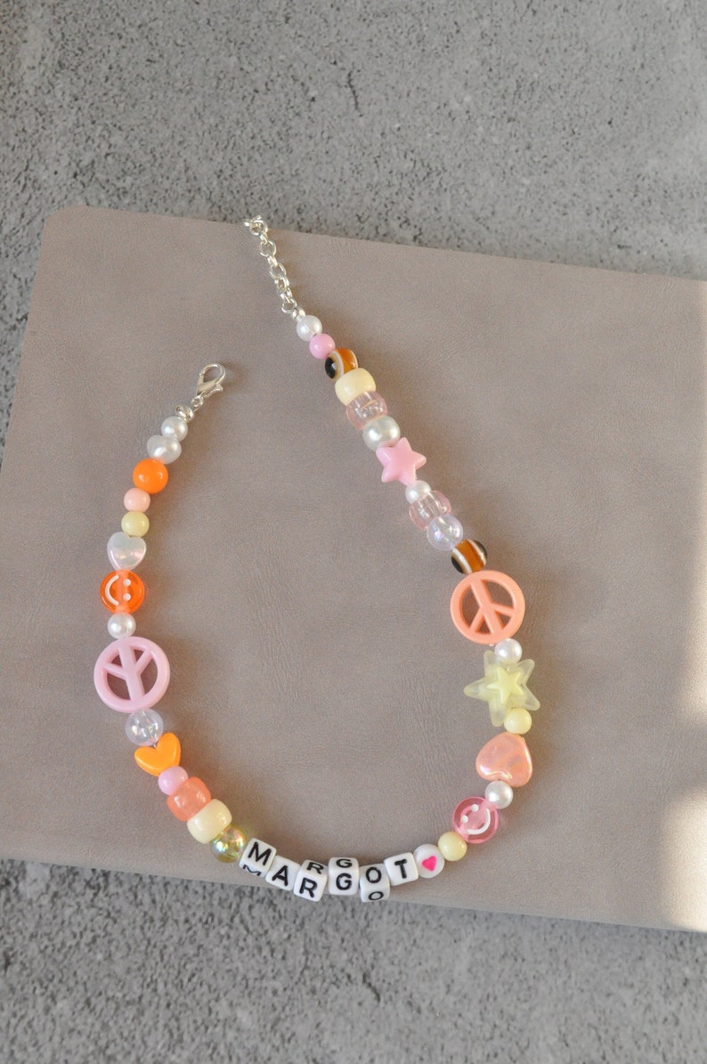 May include: A colorful beaded necklace with pink peace signs, yellow stars, orange hearts, and white pearl beads. The necklace also features the name "MARGOT" spelled out in white letter beads.