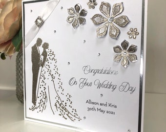 May include: A white wedding card with a silver silhouette of a bride and groom. The card is decorated with silver glitter flowers and rhinestones. The text on the card reads "Congratulations on your Wedding Day, Allison and Kris, 30th May 2021".