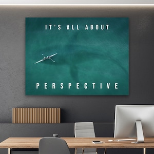 May include: A framed teal artwork with the text "IT'S ALL ABOUT PERSPECTIVE" in white. The image depicts a rowing boat on the water. The artwork is displayed on a dark gray wall above a wooden desk.