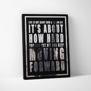 May include: Black framed canvas art with the quote "LIFE IS NOT ABOUT HOW HARD YOU HIT IT'S ABOUT HOW HARD YOU CAN GET HIT AND KEEP MOVING FORWARD" in white and gray lettering. The background is a blurred image.