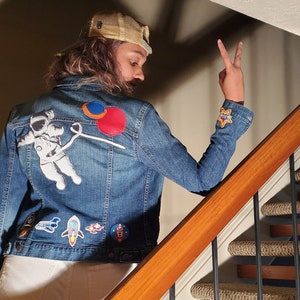 May include: A blue denim jacket with various patches, including an astronaut, a rocket, and a planet. The jacket is worn by a person with long brown hair and a beige baseball cap.