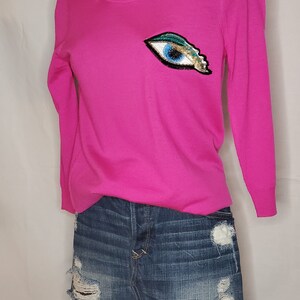 May include: A pink long-sleeved sweater with a blue eye sequin patch on the front. The sweater is paired with a denim mini skirt with distressed details.
