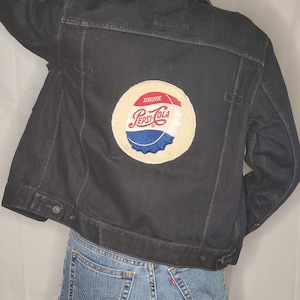 May include: A black denim jacket with a vintage Pepsi-Cola bottle cap patch on the back. The patch is white with red and blue accents and says "Drink Pepsi-Cola".