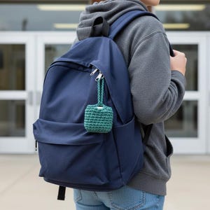 May include: A navy blue backpack with a small, green crocheted pouch attached to the zipper. The backpack has a front pocket and padded shoulder straps. The pouch is a small, square shape with a drawstring closure. The background shows a brick building and glass doors.