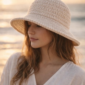 May include: A cream-colored, crocheted sun hat with a wide brim. The hat is made of a textured yarn and has a slightly rounded crown. The hat is worn by a person with long, wavy auburn hair and a white top.