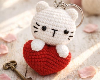 Puede incluir: Un llavero de gato blanco hecho a mano con ojos y bigotes negros, sosteniendo un corazón rojo. El llavero tiene un anillo plateado y es un accesorio encantador y caprichoso. La cara del gato tiene una forma de "T" negra.