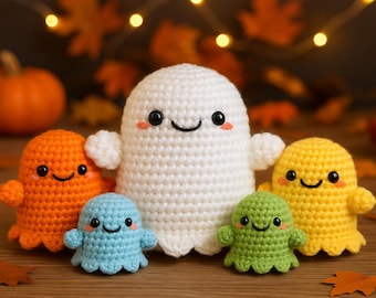 May include: A collection of five crocheted ghost figures in various colors: white, orange, blue, green, and yellow. Each ghost has a smiling face with black eyes and rosy cheeks. The ghosts are arranged on a wooden surface, with autumn leaves and a pumpkin in the background.