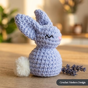 Puede incluir: Un conejito de ganchillo azul claro con una cola de pompón blanca, ojos cosidos negros y una nariz rosa. El conejito está sobre una superficie de madera. El texto "Crochet Modern Design" está en la esquina inferior derecha.