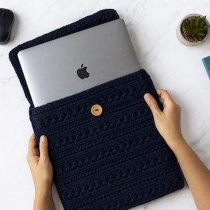 May include: A navy blue, crocheted laptop sleeve with a textured pattern and a wooden button closure. The sleeve holds a silver laptop with the Apple logo. The sleeve is designed to protect a laptop.