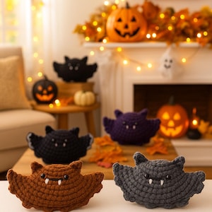 Op de afbeelding: Een verzameling handgemaakte gehaakte Halloween-decoraties, waaronder vleermuizen en pompoenen. De vleermuizen zijn in verschillende kleuren: bruin, zwart, paars en grijs. De pompoenen zijn oranje met uitgesneden gezichten.