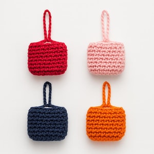 Peut inclure: Quatre petits objets carrés en crochet, rouge, rose, bleu marine et orange. Chacun a une boucle en haut pour suspendre. Les objets sont espacés uniformément sur un fond blanc. Le point de crochet est visible.