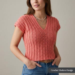 Peut inclure: Un haut en crochet corail à manches courtes et col en V. Le haut présente un motif texturé et ajouré, porté avec un jean bleu et une ceinture marron. Le texte "Crochet Modern Design" est visible en bas à droite.