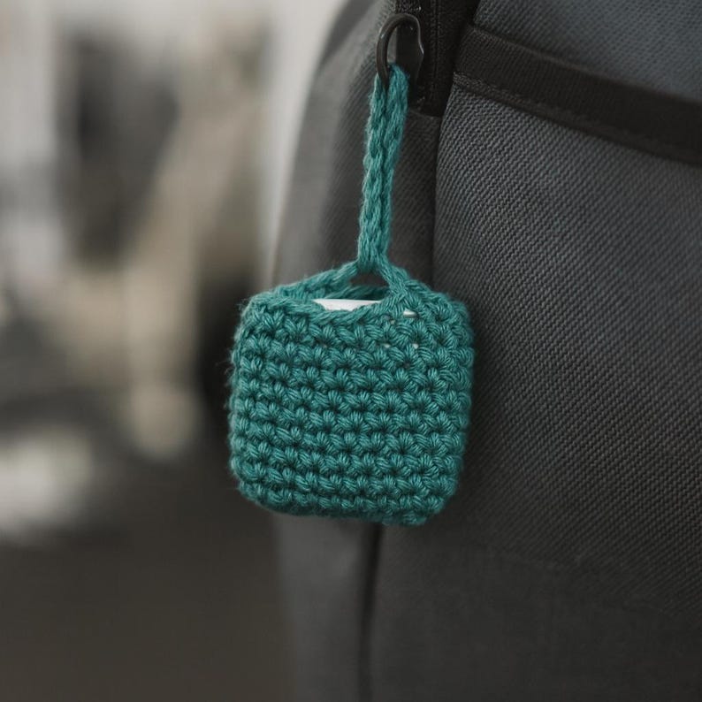 May include: A teal, square-shaped crochet case with a loop for hanging. The case is attached to a black zipper. The crochet has a tight, textured stitch. The case appears to be designed to hold a small electronic device.