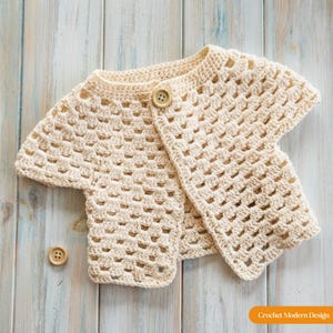 Peut inclure: Cardigan en crochet de couleur crème avec des manches courtes et une fermeture à bouton unique. Le motif ajouré est visible, et un bouton de rechange est placé en dessous. Le vêtement est présenté sur une surface en bois bleu clair et blanc.