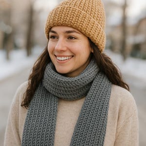 May include: A woman wearing a tan knit beanie, a gray knit scarf, and a beige sweater. The scarf is wrapped around the neck and has a textured pattern. The background is blurred, with trees and a snowy path visible.