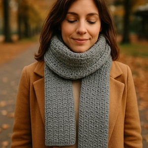 May include: A gray knitted scarf with a textured pattern is draped around the neck. The scarf is worn over a tan coat. The background is blurred, with trees and foliage in autumn colors.