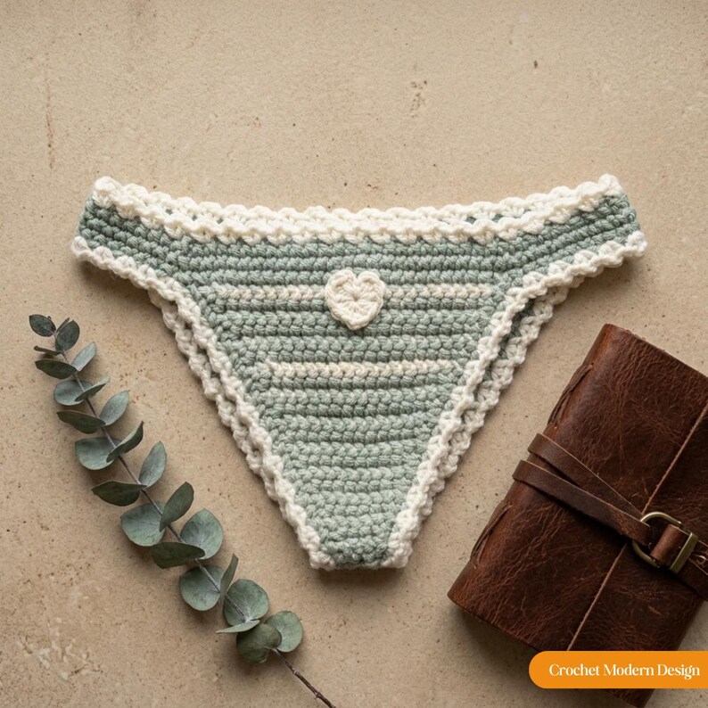 May include: A sage green crochet bikini bottom featuring a white scalloped trim along the edges, a small white heart applique in the centre, and horizontal white stripes. The text "Crochet Modern Design" is visible in the bottom right corner.