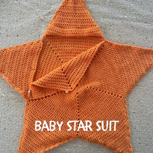 Può includere: Una tutina per neonati a forma di stella all'uncinetto di colore arancione, dotata di cappuccio e chiusura con bottone. Il capo presenta un motivo a maglia testurizzato e la scritta bianca BABY STAR SUIT sulla parte anteriore.
