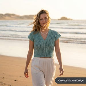 May include: A turquoise crochet top with a V-neck and short sleeves. The top is paired with white pants. The image is taken on a beach with the ocean in the background. The text "Crochet Modern Design" is in the bottom right corner.