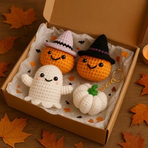 Peut inclure: Un ensemble de décorations d'Halloween faites à la main au crochet, présentées dans une boîte en carton. L'ensemble comprend deux citrouilles oranges avec des chapeaux de sorcière, un fantôme blanc et un porte-clés citrouille blanche. Les citrouilles ont des visages souriants.
