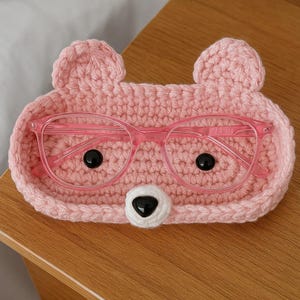 Peut inclure: Un plateau rose en forme d'ours en crochet contenant une paire de lunettes roses. Le plateau a deux petites oreilles et un nez blanc avec des yeux noirs. Les lunettes ont une monture transparente et reposent à l'intérieur du plateau.