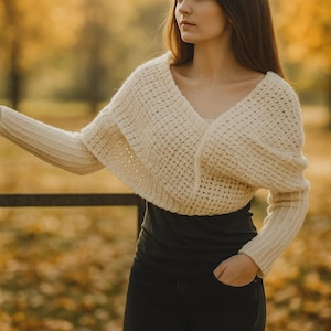 Peut inclure: Boléro en tricot crème à devant ouvert et manches longues. Le boléro présente un col en V profond et un motif en maille ajourée texturée. Le modèle porte un haut noir et un pantalon noir. L'arrière-plan est flou avec des couleurs d'automne.
