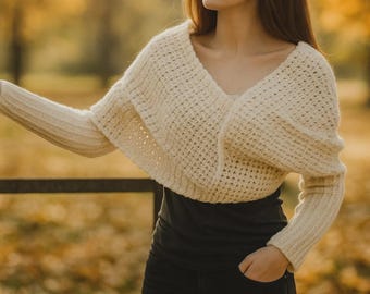 May include: Cream-colored, open-front knitted shrug with long sleeves. The shrug features a deep V-neck and a textured, open weave design. The model is wearing a black top and black pants. The background is blurred with autumn colors.