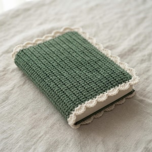 May include: A rectangular book cover made of dark green crocheted yarn with a textured ribbed pattern. The cover features a decorative cream-coloured scalloped crochet border that frames the entire edge of the book, providing a finished look.