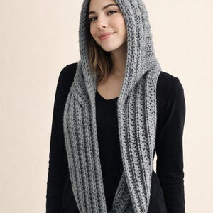 Puede incluir: Bufanda con capucha gris hecha de material de punto. La bufanda es larga y se envuelve alrededor del cuello, con la capucha levantada. La bufanda es de color gris liso y parece hecha a mano.