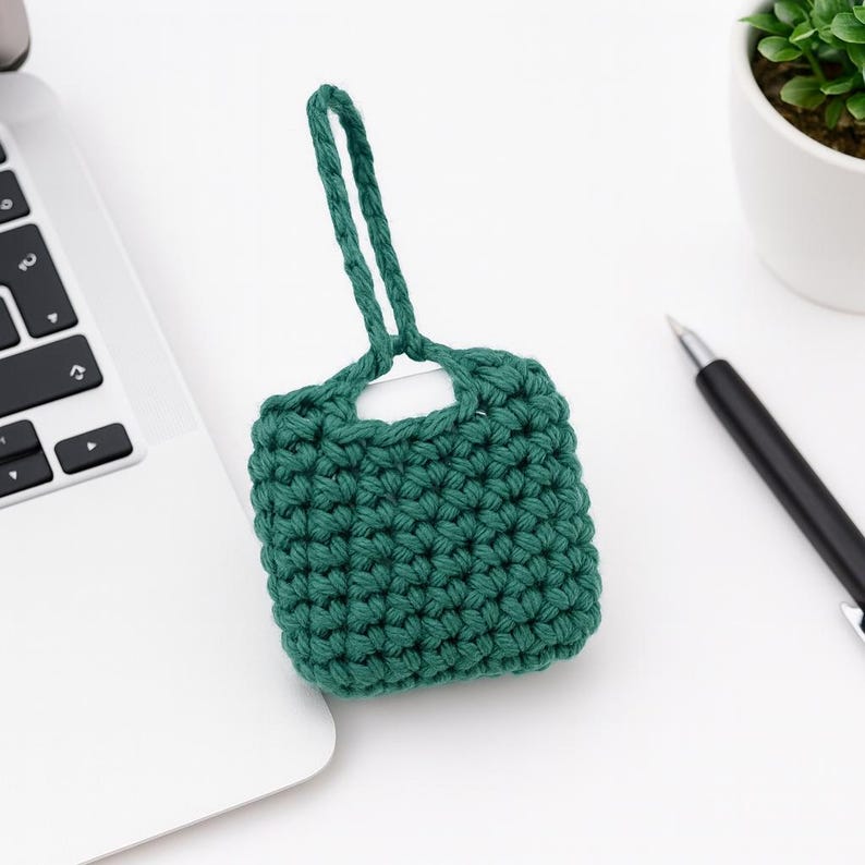 May include: A handcrafted, forest green crochet case with a loop handle. The case is square-shaped and sits on a white surface next to a laptop keyboard, a pen, and a small potted plant. The case is made of a thick, textured yarn.