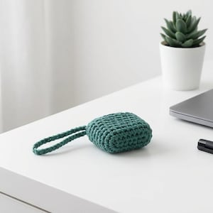 May include: A teal, crocheted square case with a matching loop handle rests on a white surface. A small succulent in a white pot, a closed laptop, and a black pen are also on the surface.