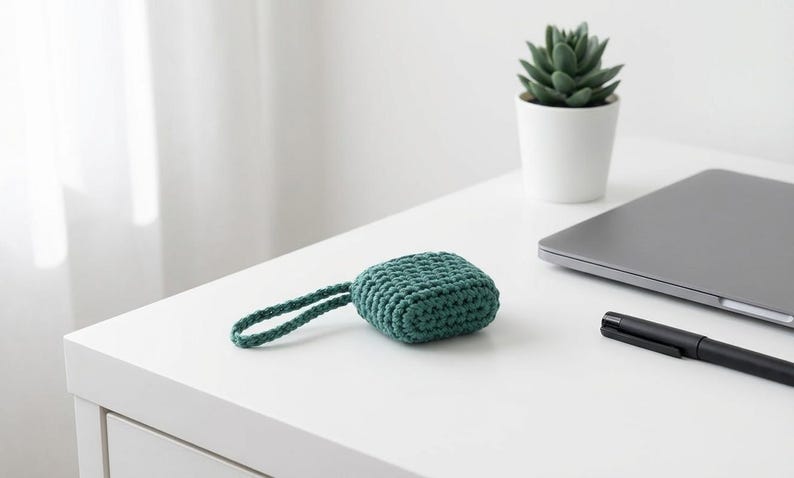 May include: A teal, crocheted square case with a matching loop handle rests on a white surface. A small succulent in a white pot, a closed laptop, and a black pen are also on the surface.