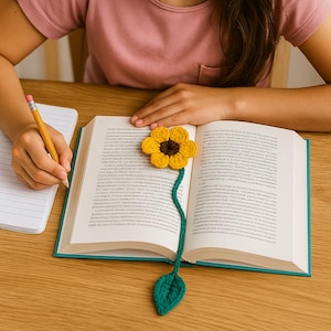 Op de afbeelding: Een gehaakte zonnebloem bladwijzer met een gele bloem, bruin midden, groene stengel en blad. De bladwijzer ligt in een open boek met witte pagina's en een turquoise kaft. Een potlood en notitieboek zijn ook zichtbaar.