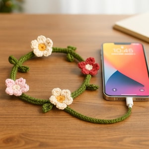 Peut inclure: Un organiseur de câble en crochet vert en forme de couronne florale, avec des fleurs crème, roses et rouges. Un smartphone est connecté à l'organisateur. L'écran du téléphone affiche l'heure 10h46. L'organisateur est sur une surface en bois.