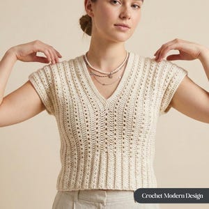 Peut inclure: Haut en crochet crème à manches courtes et col en V. Le haut présente un motif texturé et ajouré. Un collier à plusieurs rangs est également visible. Le texte "Crochet Modern Design" est en bas de l'image.