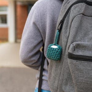 May include: A teal crocheted case with a small handle is attached to a gray backpack. The case holds a white, rounded object, likely earbuds or a similar small electronic device. The crochet work has a textured appearance.