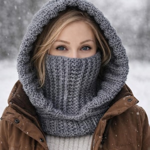 Peut inclure: Un ensemble cagoule et col tricoté gris. La cagoule est relevée, couvrant la tête et le cou, le col couvrant le bas du visage. La texture tricotée est visible, et la couleur est un gris mat. La personne porte une veste marron.