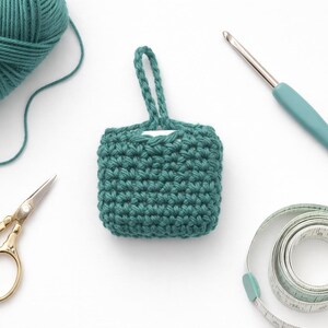 May include: An overhead shot of crochet supplies on a white surface. A teal yarn ball, a small teal crocheted square with a handle, gold scissors, a teal crochet hook, and a measuring tape are visible.