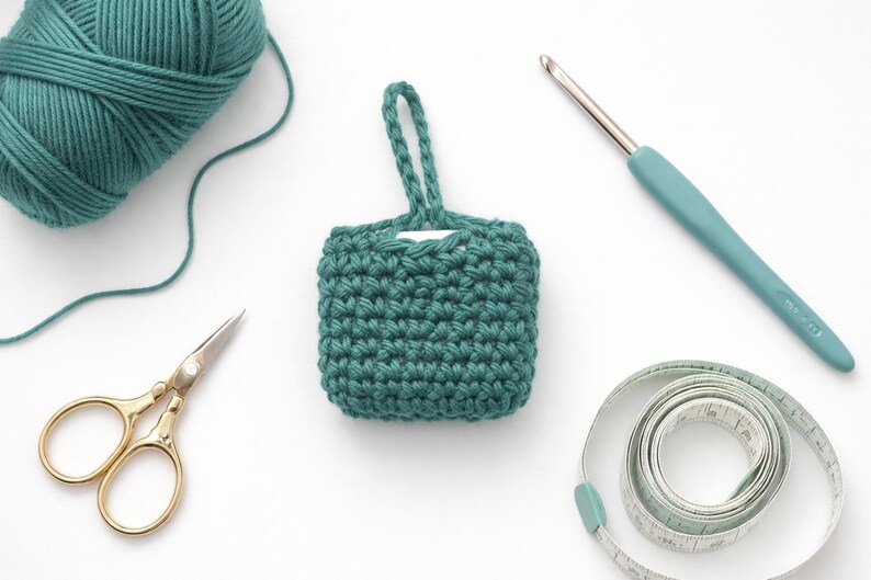 May include: An overhead shot of crochet supplies on a white surface. A teal yarn ball, a small teal crocheted square with a handle, gold scissors, a teal crochet hook, and a measuring tape are visible.