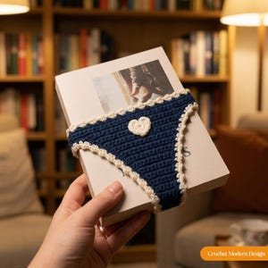 May include: A dark blue crochet book sleeve featuring a white heart appliqu&eacute; in the centre and a decorative white scalloped border along the edges. The sleeve is fitted around a rectangular book with a white cover and a printed image on the front.