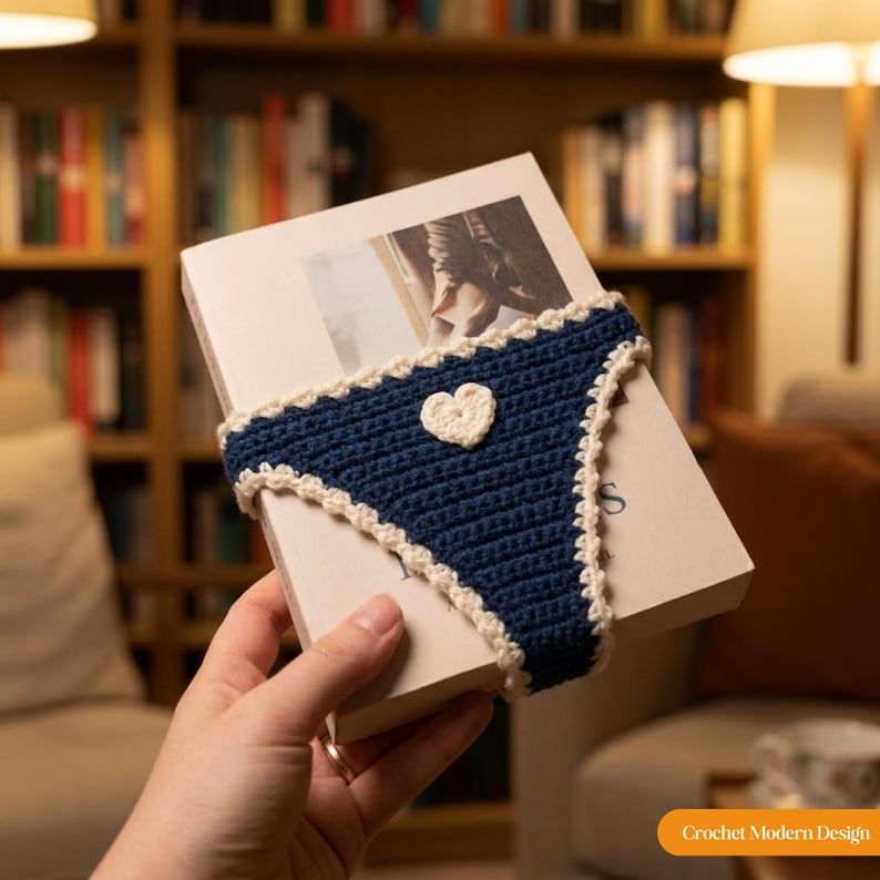 May include: A dark blue crochet book sleeve featuring a white heart appliqu&eacute; in the centre and a decorative white scalloped border along the edges. The sleeve is fitted around a rectangular book with a white cover and a printed image on the front.